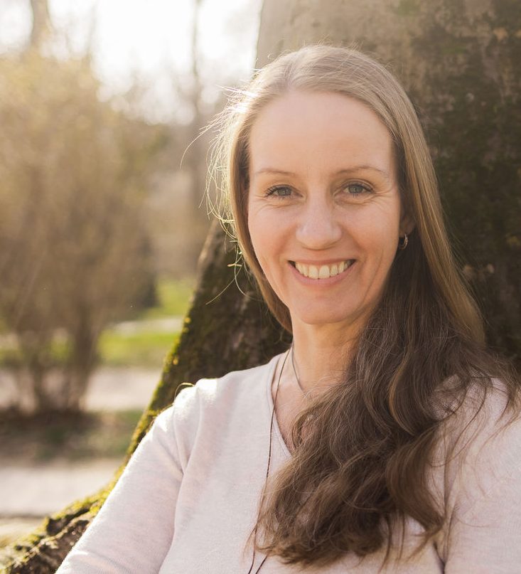 Pia Messerer-Heilpraktikerin für Psychotherapie Lächelnde Frau mit langen Haaren, sitzt an einem Baum im Sonnenlicht.